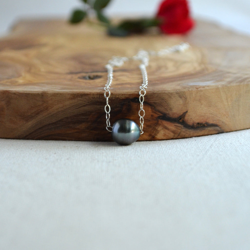 A silver chain necklace with a single dark gray pearl pendant, displayed on a rustic wooden board with a blurred red rose in the background.