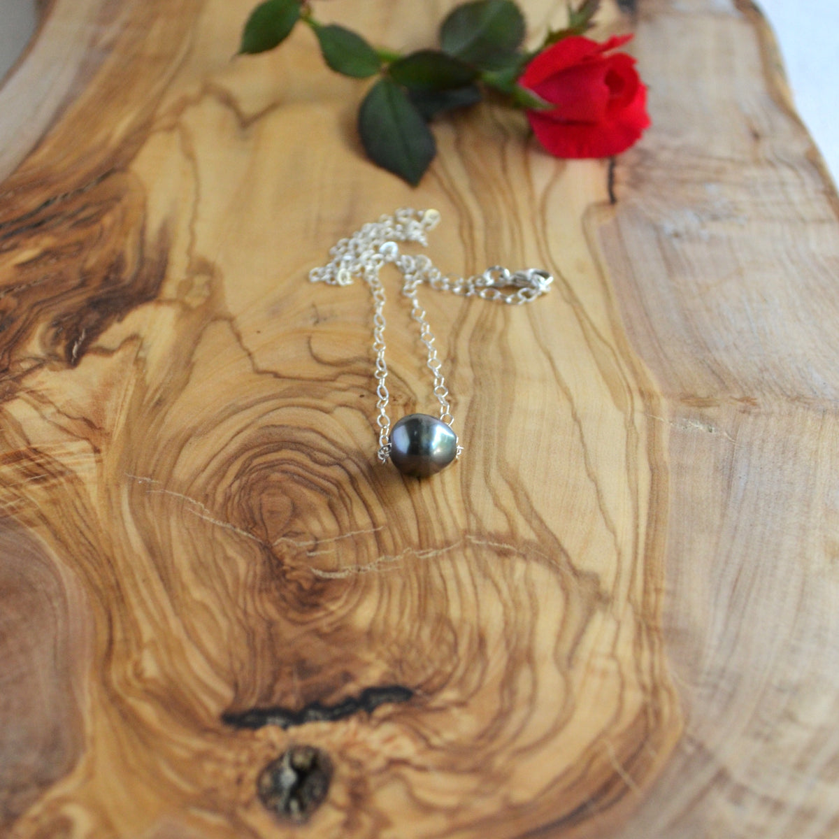 A silver chain necklace with a dark gray-black round pearl pendant, displayed on a rustic olive wood slice with a red rose in the background.