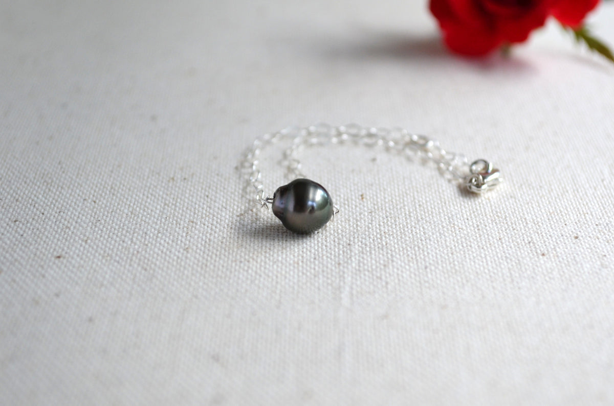 A delicate silver bracelet featuring a single dark gray baroque pearl, displayed on a textured off-white fabric background with a blurred red rose in the upper right corner.