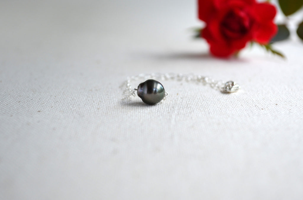 A delicate silver chain bracelet featuring a single Tahitian pearl, resting on a textured off-white fabric surface with a blurred red rose in the background.