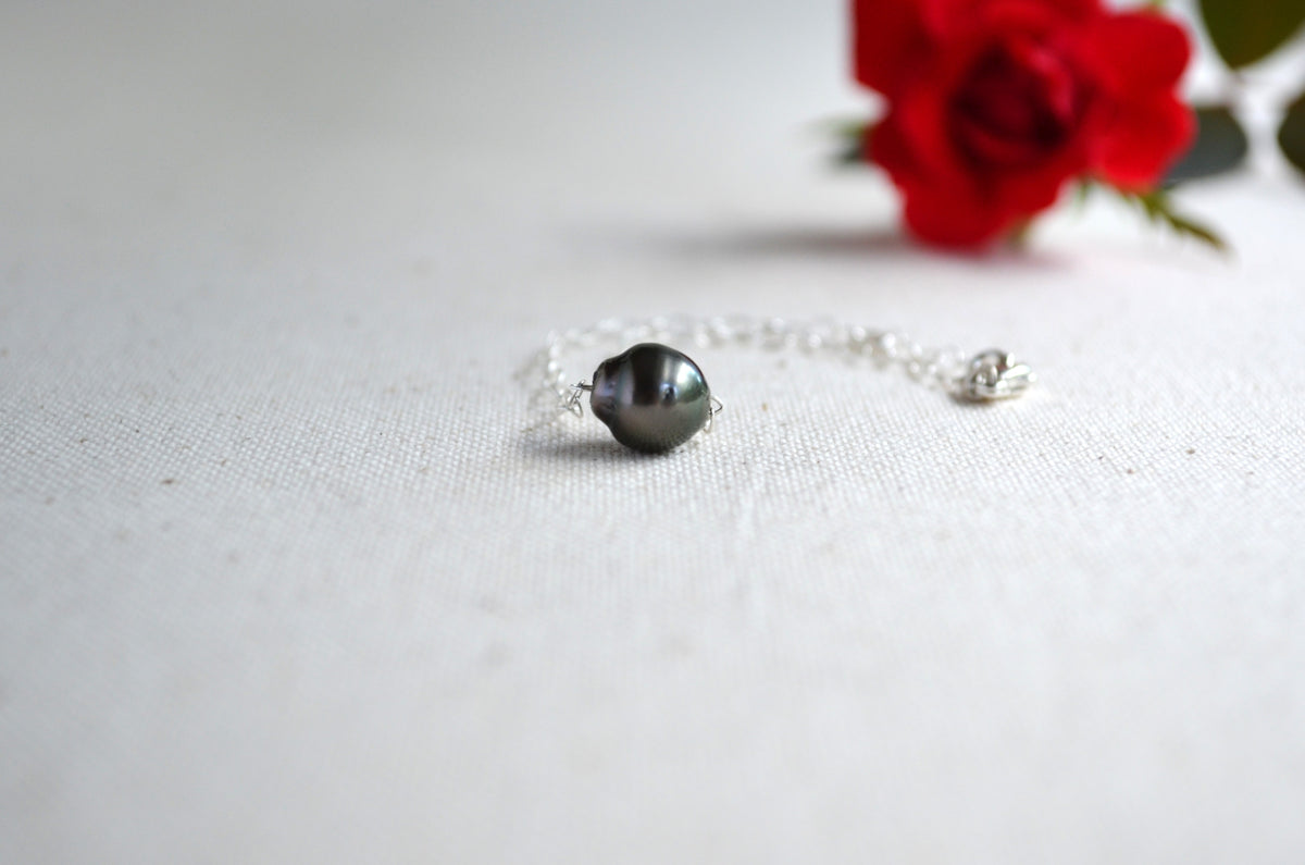 A delicate silver chain bracelet featuring a single Tahitian pearl, resting on a textured off-white fabric surface with a blurred red rose in the background.
