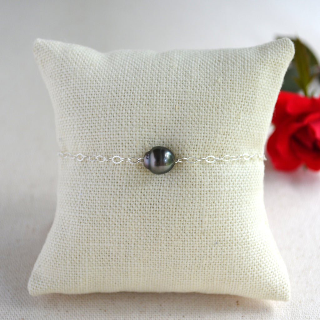 A delicate silver chain bracelet with a single dark gray Tahitian pearl bead, displayed on a cream-colored textured fabric pillow with a red rose in the background.