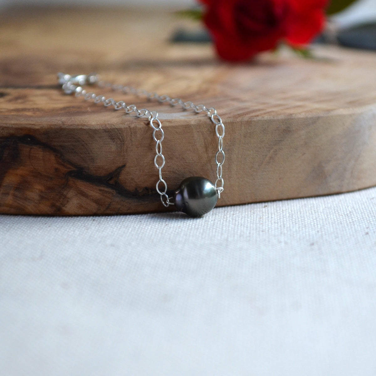 A silver chain bracelet with a single dark gray pearl pendant, resting on a rustic wooden board with a blurred red rose in the background.