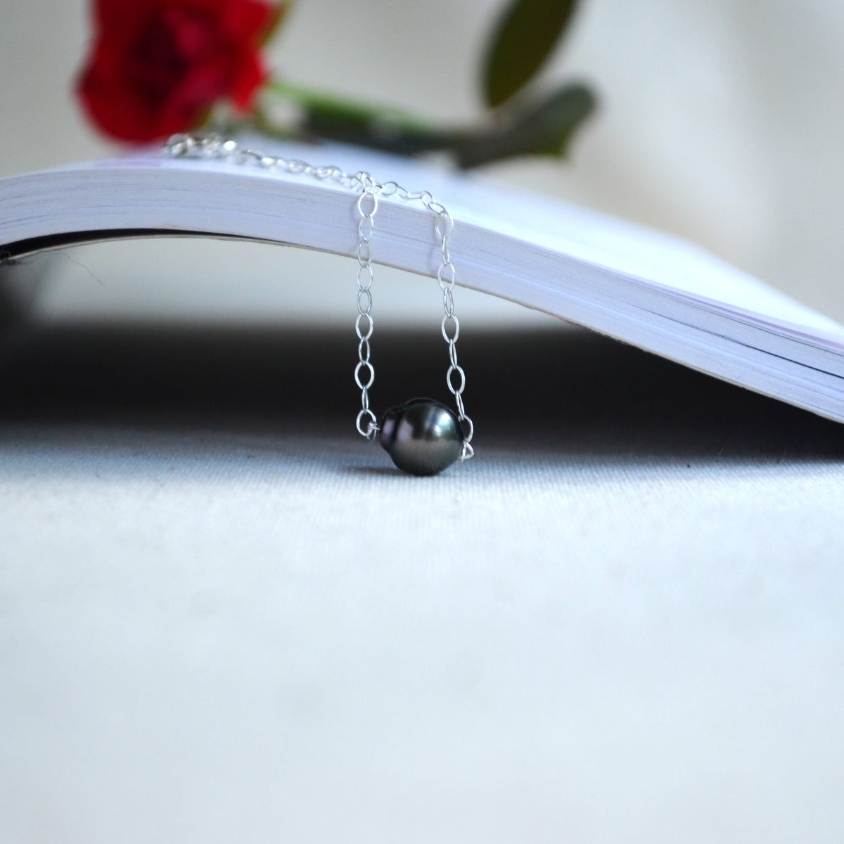 A delicate silver chain bracelet with a single dark gray pearl pendant, draped over the edge of an open book with a blurred red rose in the background.