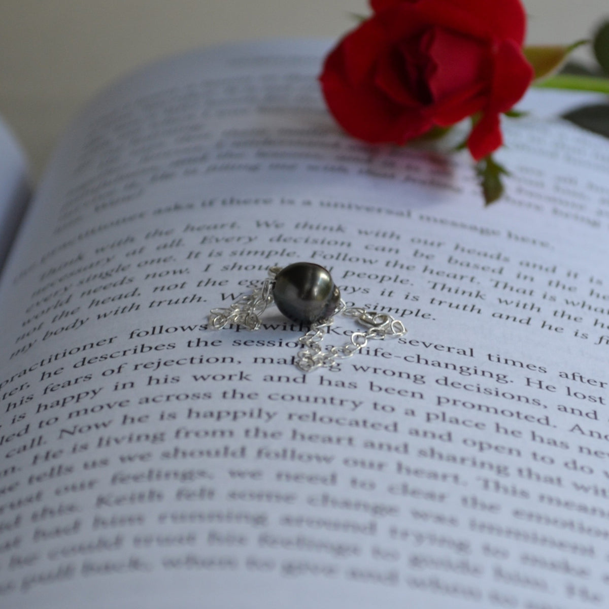Sterling silver chain Tahitian pearl bracelet and delicate floral scrollwork, resting on an open book with a red rose in the background.