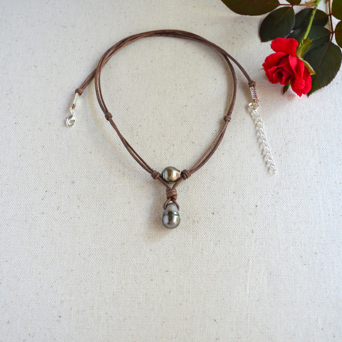 A brown leather cord necklace with a dark gray baroque Tahitian pearls pendant, and a silver chain extender, displayed on a textured cream fabric next to a red rose.