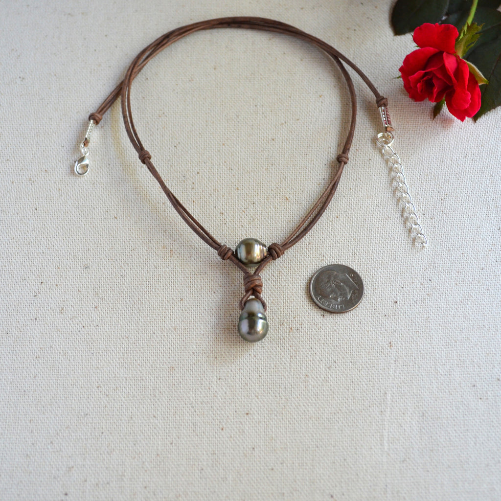 A brown leather cord necklace with a central Tahitian pearls, and a silver-toned chain extender, displayed on a beige fabric surface next to a coin for scale and a red flower.