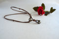 A dark brown cord necklace with two dark gray teardrop-shaped pearls, one larger and one smaller, displayed on a light beige textured surface with a red rose in the background.