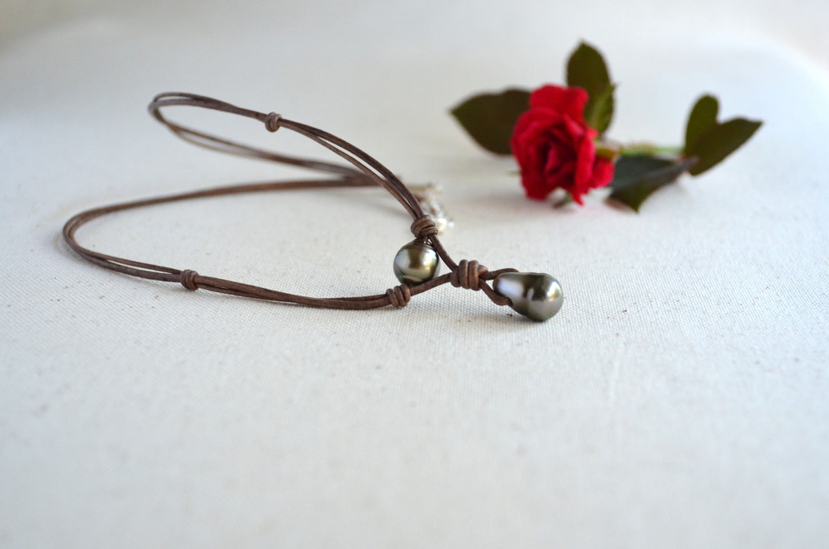 A brown cord bracelet with two dark Tahitian pearls, displayed on a textured white surface next to a small red rose.