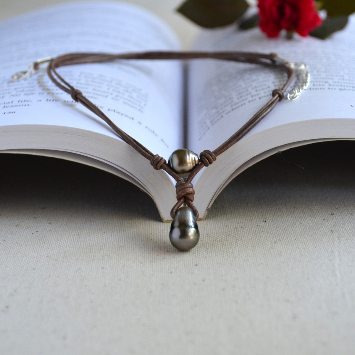 A brown leather cord necklace with a dark gray teardrop pearl pendant and a metallic bronze bead, displayed on an open book with a red rose in the background.