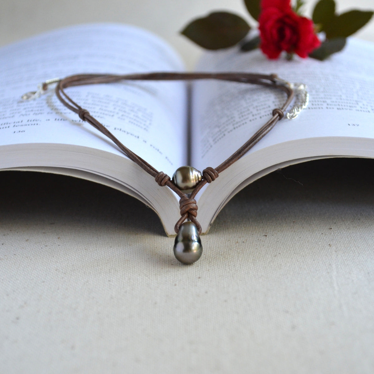 A brown leather cord necklace with two Tahitian pearls, displayed on the pages of an open book with a red rose in the background.