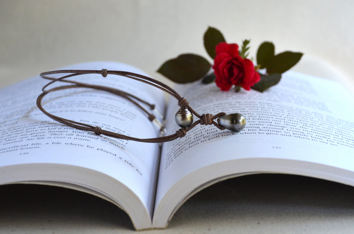 A brown leather necklace with two Tahitian pearls, resting on an open book with a red rose.