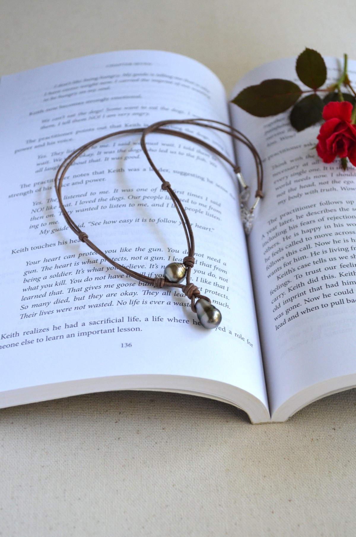 Leather necklace with two Tahitian pearls draped over the pages of an open book with a red rose resting on the right page.