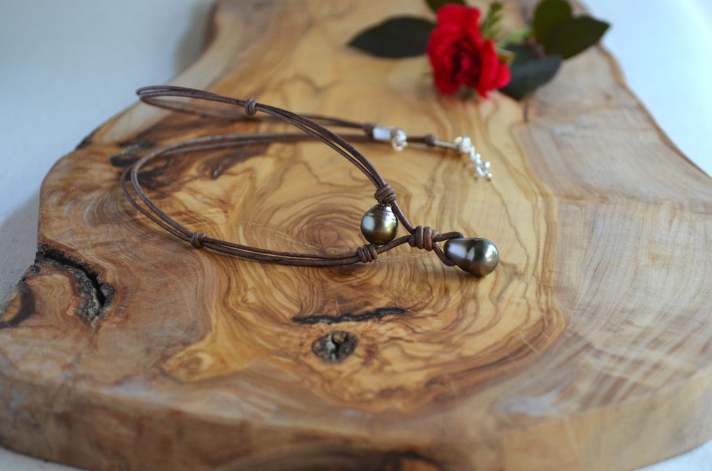 A dark brown leather necklace with two Tahitian pearls, displayed on a rustic wood slice with a red rose in the background.