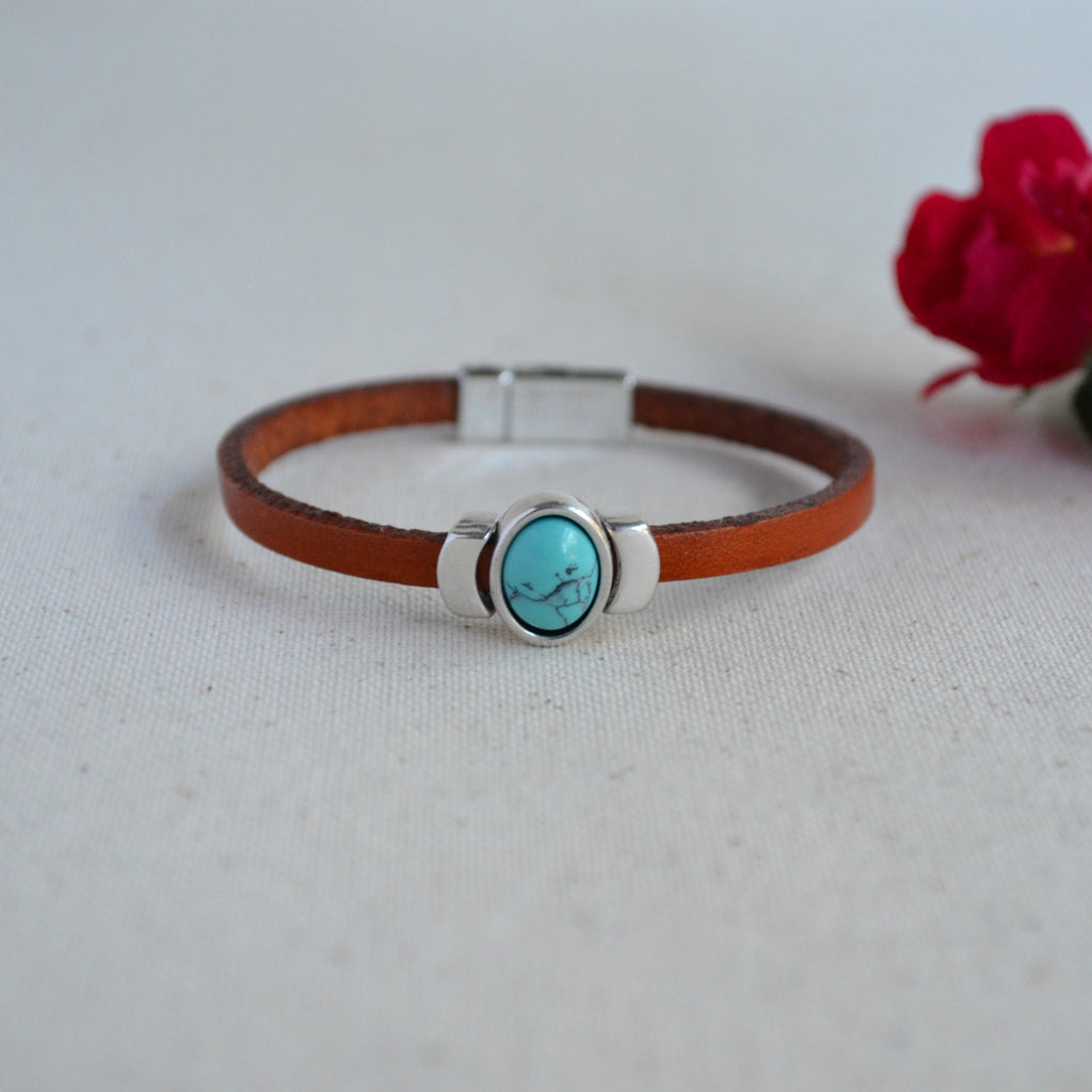 A brown leather bracelet with a silver-toned magnetic clasp and a central oval turquoise stone bezel, displayed on a light textured fabric background with a blurred red rose in the background.
