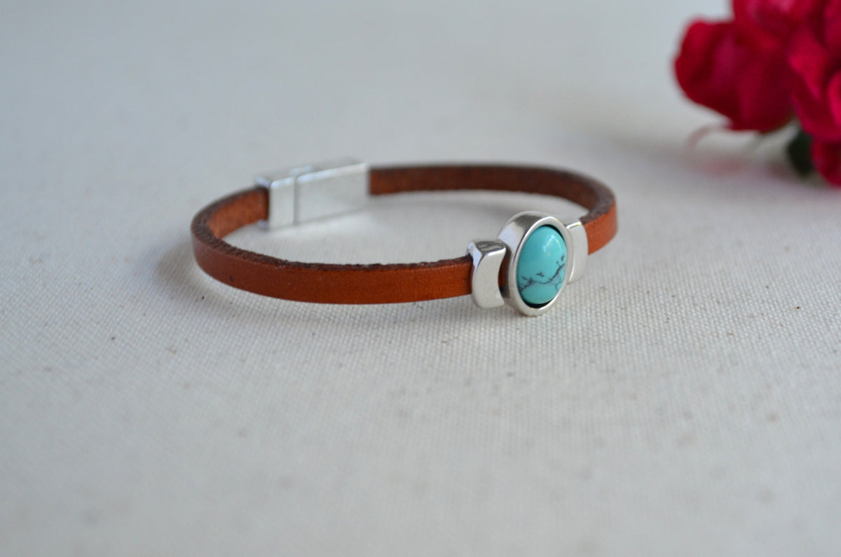 A brown leather bracelet with a silver-toned magnetic clasp and a central oval turquoise stone bezel setting, resting on a light beige textured surface with a blurred red flower in the background.