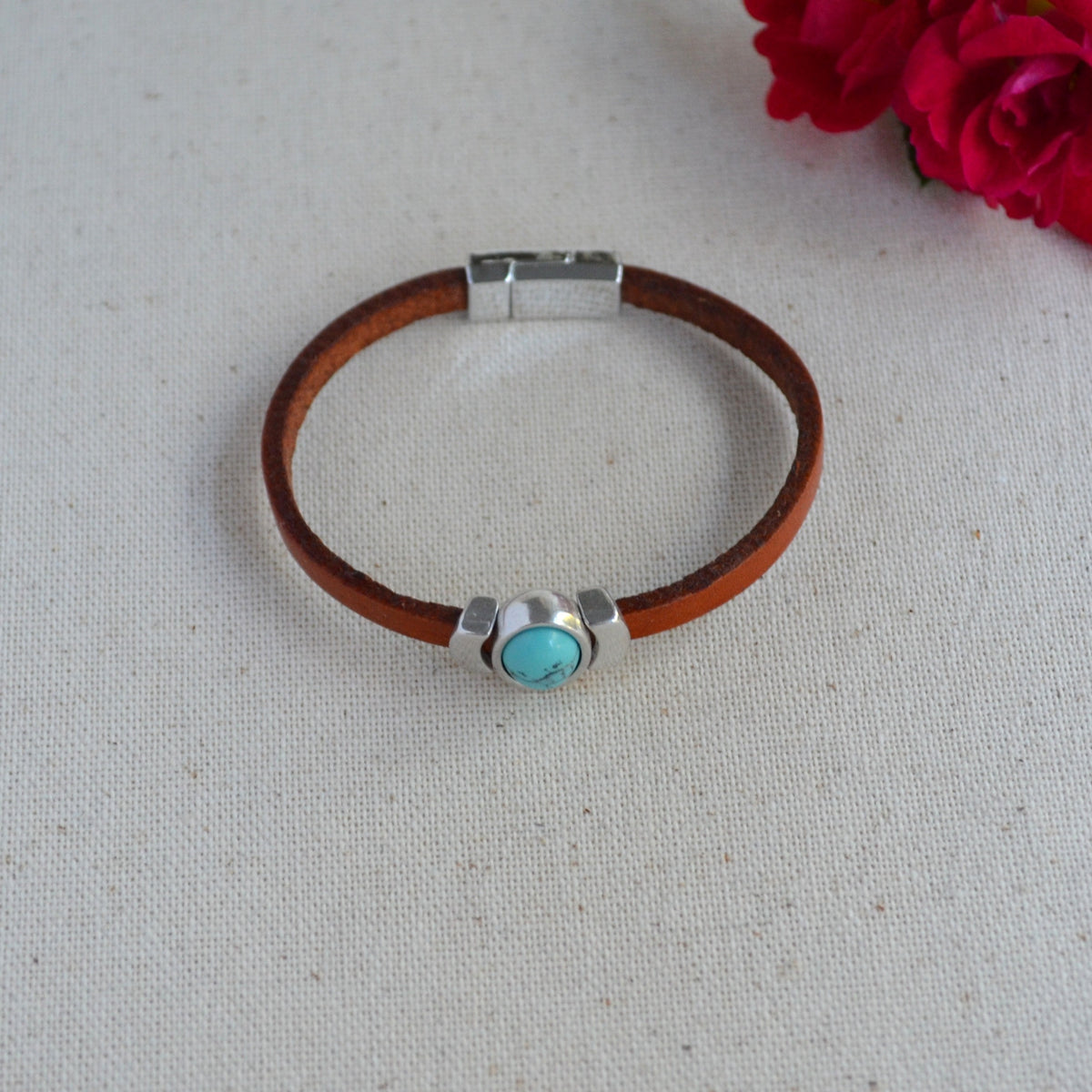 A brown leather bracelet with a silver-toned magnetic clasp and a central turquoise stone bezel setting, displayed on a textured off-white fabric background with red roses in the corner.