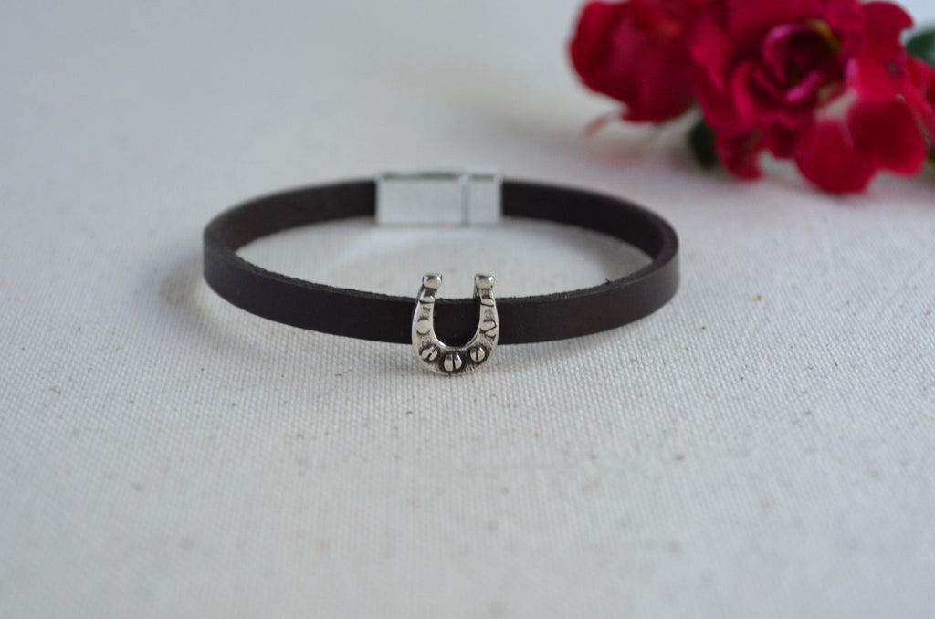 A dark brown leather bangle bracelet with a silver horseshoe charm, displayed on a textured white fabric background with red roses in the background.