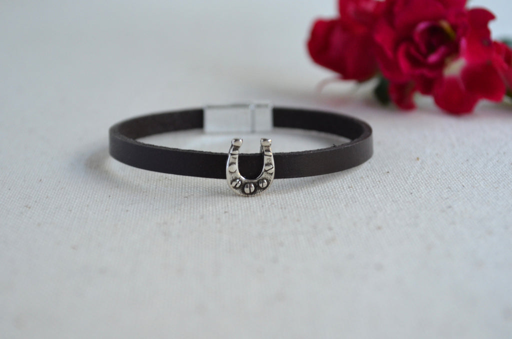 A brown leather bangle bracelet with a silver horseshoe charm embellished with small rhinestones, resting on a light textured surface with blurred red flowers in the background.