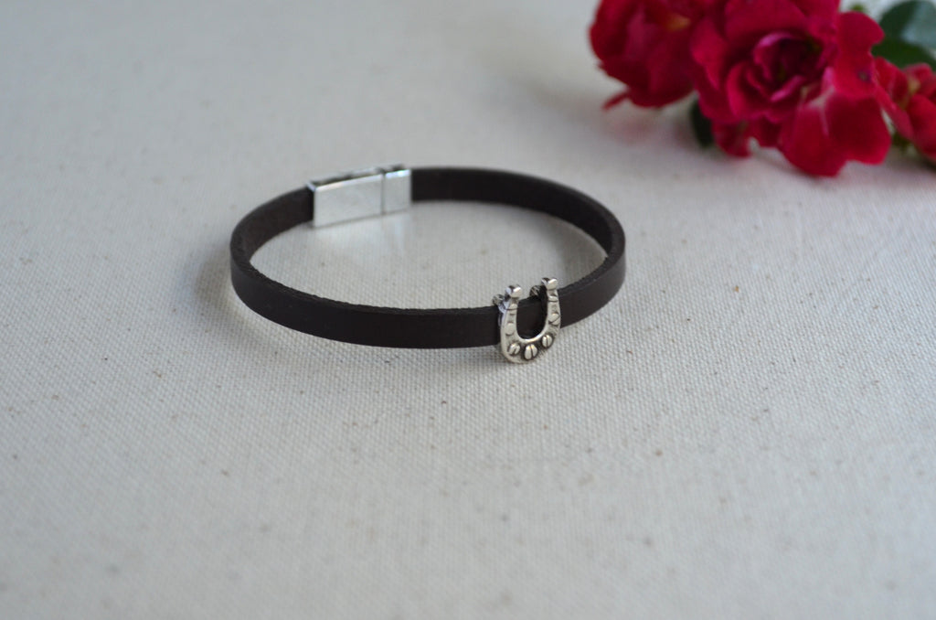 A black leather bracelet with a silver horseshoe charm embellished with small clear rhinestones, displayed on a textured light beige fabric surface with red roses in the background.