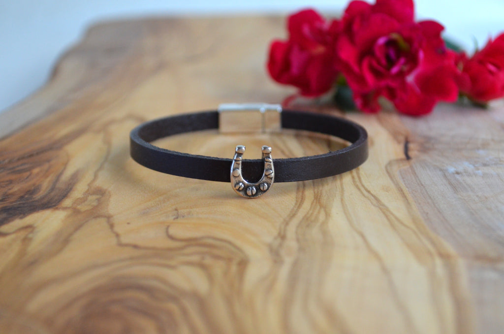 A black leather bracelet with a silver horseshoe charm, displayed on a wooden board with red flowers in the background.