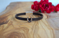 A black leather bracelet with a silver horseshoe charm, displayed on a wooden board with red flowers in the background.