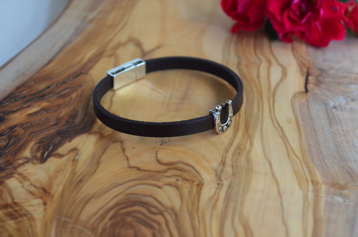 A dark brown leather bracelet with a silver horseshoe charm, displayed on a wooden board with red flowers in the background.