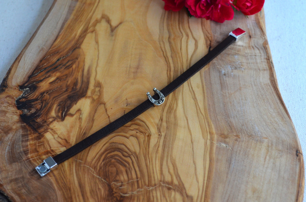A dark brown leather bracelet with silver-toned metal hardware, including a horseshoe charm and red square end caps, displayed on a rustic olive wood board with red flowers.
