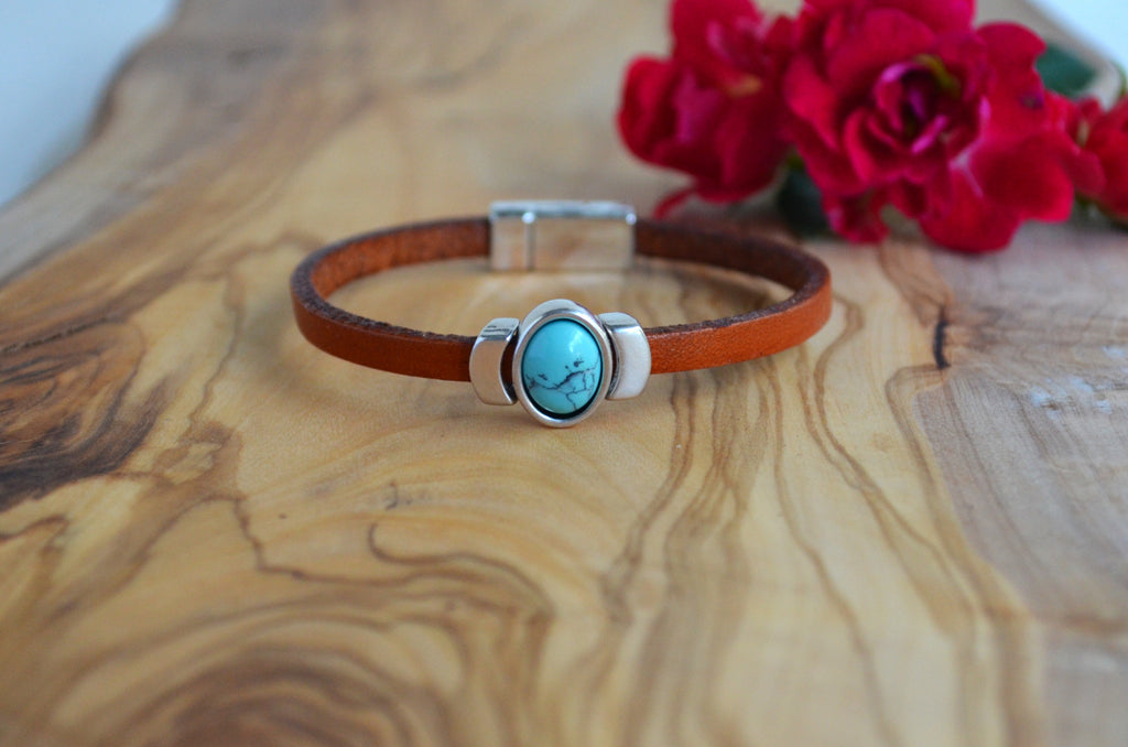A brown leather bracelet with a silver-toned clasp and a central oval turquoise stone featuring a black anchor design, displayed on a wooden surface with red flowers in the background.