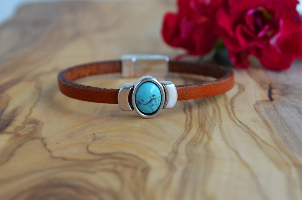 A brown leather bracelet with a silver-toned clasp and a central oval turquoise stone featuring a black bird design, displayed on a wooden surface with red flowers in the background.