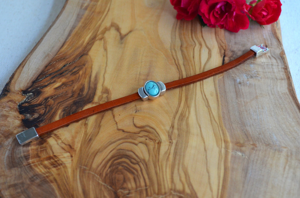 A brown leather bracelet with a silver-toned metal case and a round turquoise blue stone bezel, displayed on a rustic wood slice background with red roses in the corner.