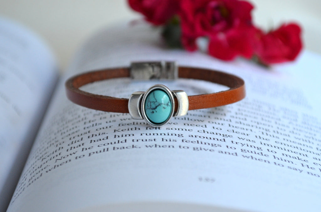 A brown leather bracelet with a silver-toned oval bezel holding a turquoise-blue stone, displayed on an open book with blurred red flowers in the background.