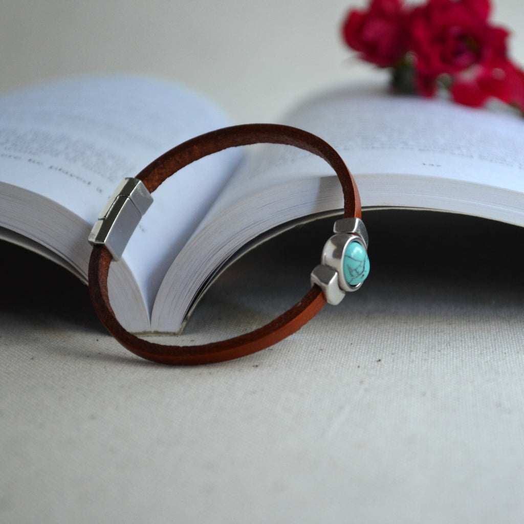 A brown leather bracelet with a silver-toned rectangular clasp and a turquoise stone accent, resting on the pages of an open book with blurred red flowers in the background.