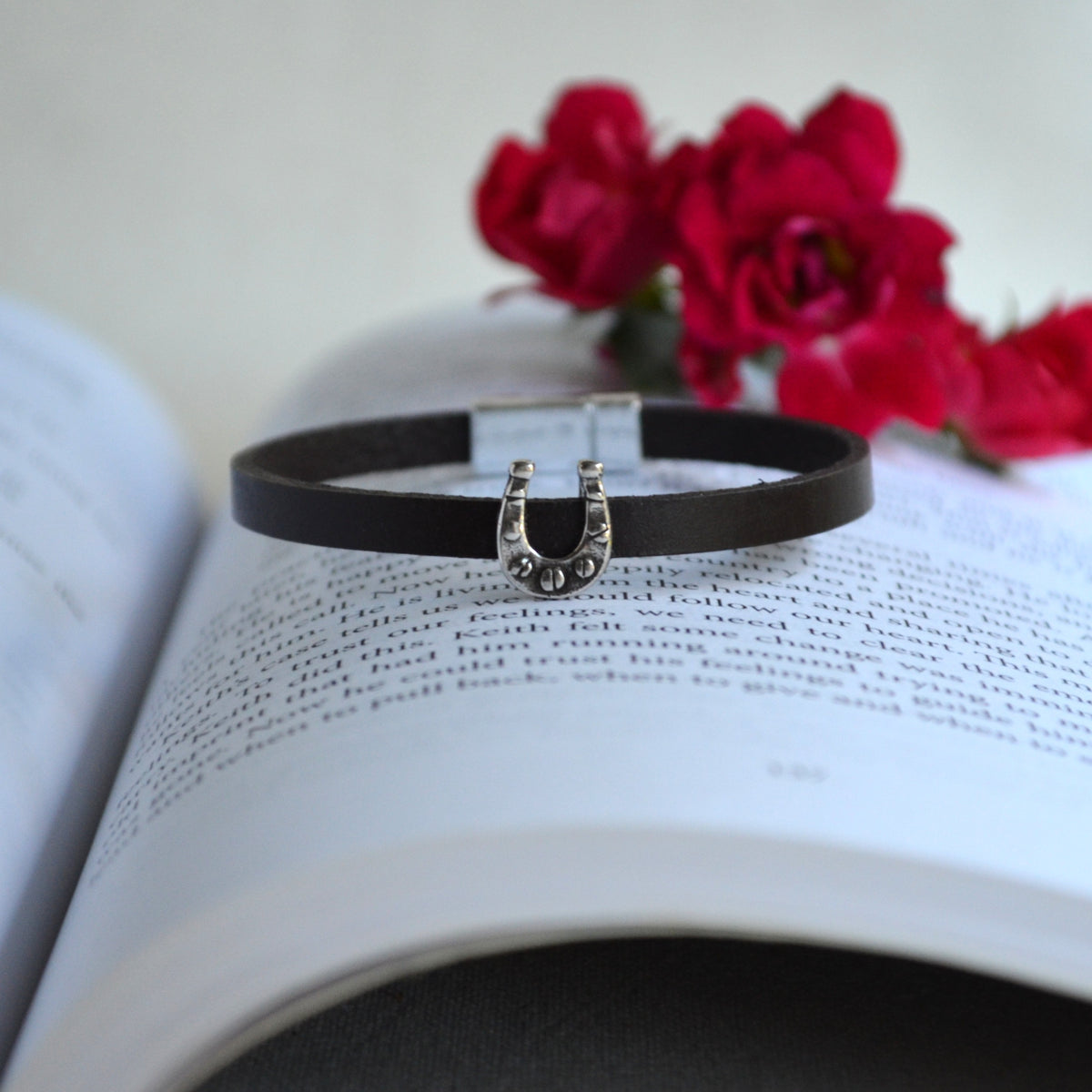 A black leather bracelet with a silver horseshoe charm embellished with small clear stones, resting on an open book with red roses in the background.