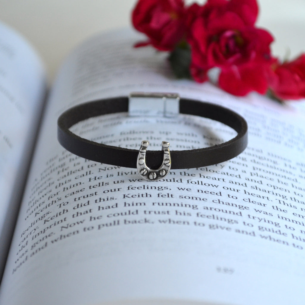 A black leather bracelet with a silver horseshoe charm, displayed on open book pages with red flowers in the background.