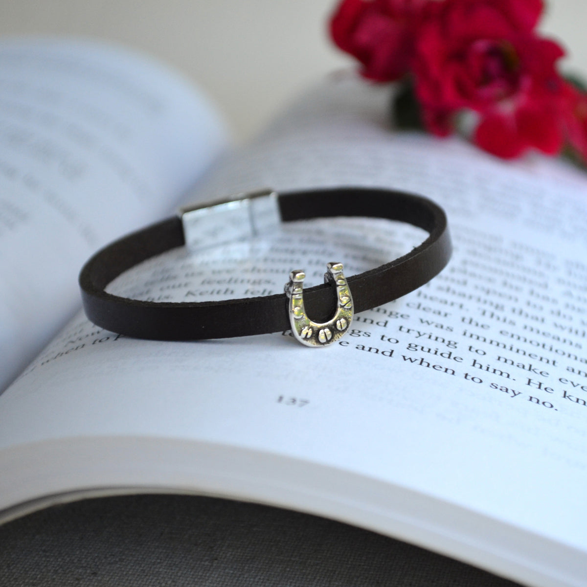 A dark brown leather bracelet with a silver horseshoe charm, resting on an open book with red roses in the background.