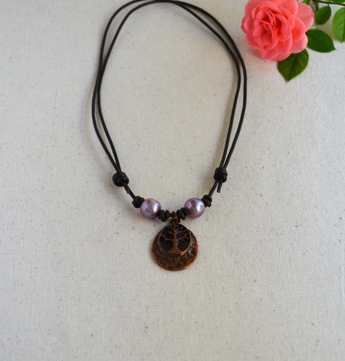 Necklace with two pink Edison pearls and Tree of life pendant on brown Greek leather cord on the light fabric background with pink rose on the back