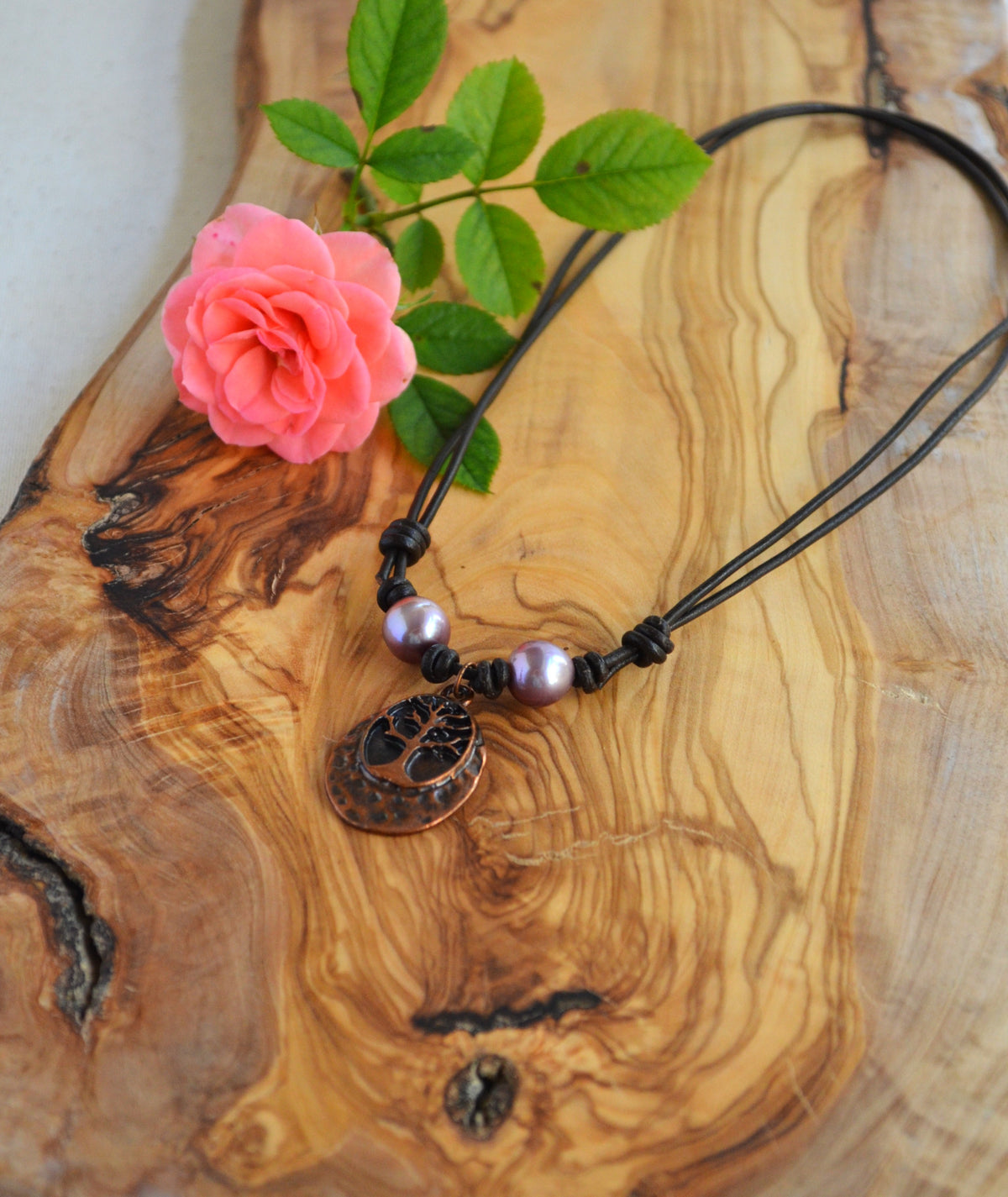 Necklace with two pink Edison pearls and Tree of life pendant on brown Greek leather cord on the wooden surface with pink rose on the left