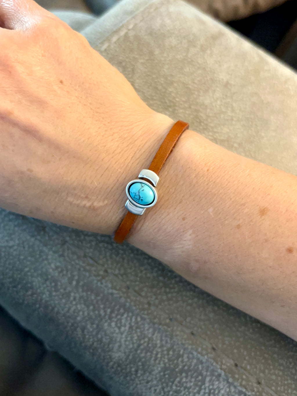 A close-up of a person's wrist wearing a slim brown leather bracelet with a round turquoise-blue stone centerpiece and a white and silver-toned metal connector.