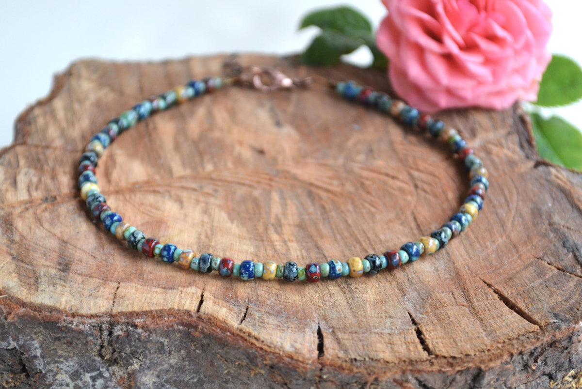 Multicolored beaded anklet bracelet on a wooden surface with a pink flower in the background