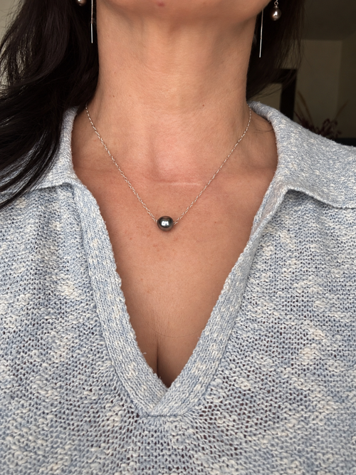A close-up of a person wearing a delicate silver chain necklace with a single dark gray round pearl pendant, paired with a light blue knit collared top.
