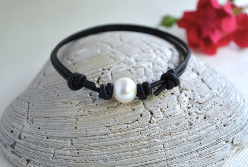 Black cord bracelet with a pearl on a textured surface with red flowers in the background