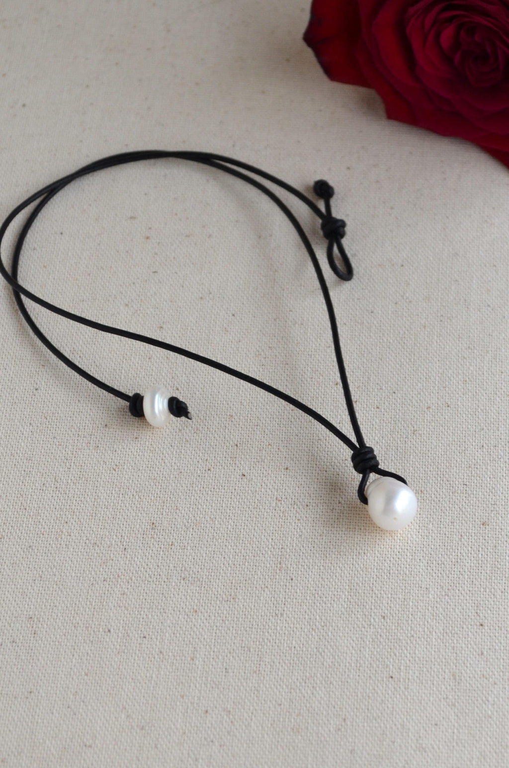 Black cord necklace with white pearls on a beige surface with a red rose.