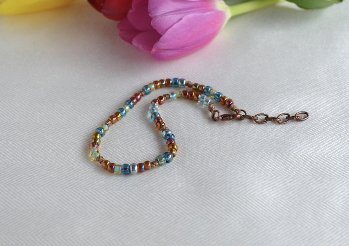 Multicolored beaded anklet on a white surface with pink flowers in the background