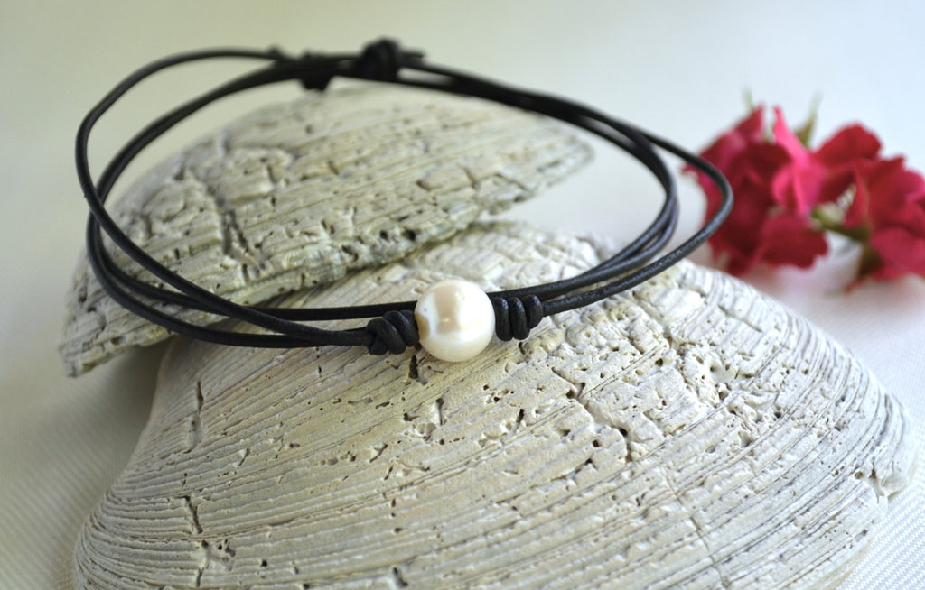 Black leather necklace with a pearl bead on a stone surface with pink flowers in the background