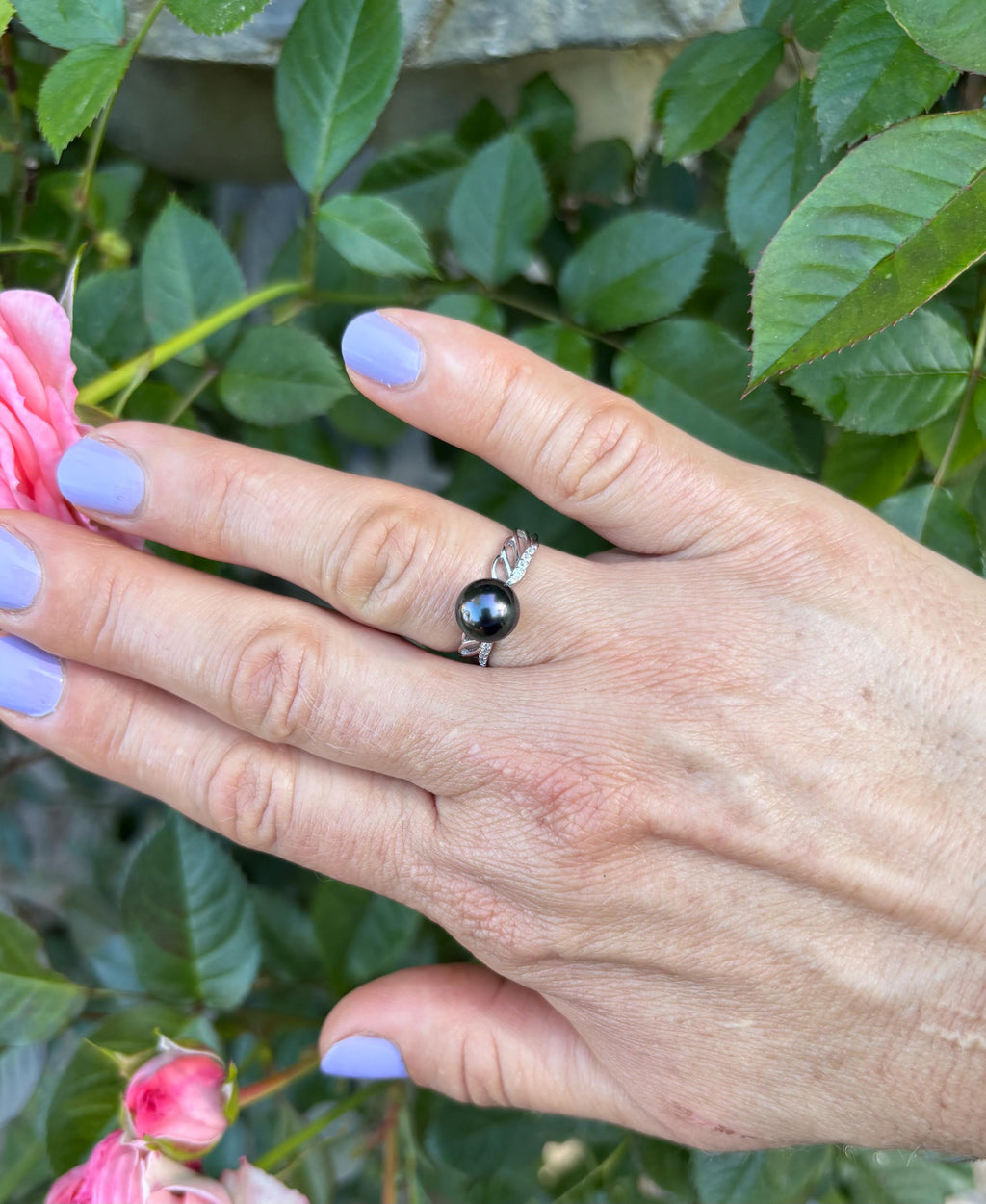 Hand wearing a black ring with a pink flower and green leaves in the background